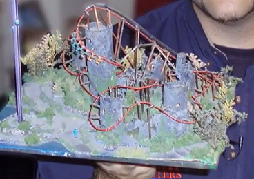 Model of a roller coaster created by and shown by a mechanical engineer named Chris Gray. He is holding a page-size board with a  handmade landscape with trees, grass, hills and the roller coaster.
